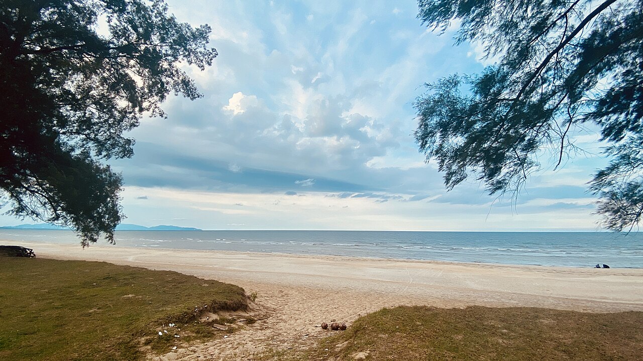 Sepat Beach in Kuantan, a wide sandy shore framed by trees under a soft blue sky